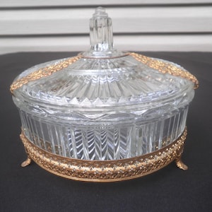 May include: A clear glass candy dish with a lid, featuring a gold-colored filigree base and rim. The dish has a ribbed design and a decorative finial on the lid. The dish is a vintage piece.