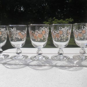 May include: Five clear wine glasses with a decorative floral pattern in gold and white. The glasses have a rounded bowl, a stem with a bulbous detail, and a circular base. The glasses are arranged in a row on a white surface.