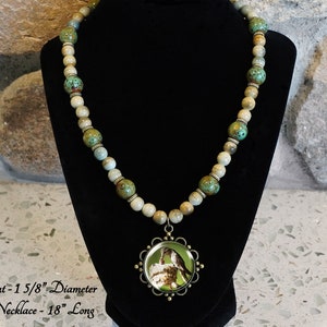 May include: A necklace with a pendant featuring a pair of birds on a green background. The necklace is made of brown and green ceramic beads and is 18 inches long. The pendant is 1 5/8 inches in diameter.  Pendant - 1 5/8" Diameter Bead Necklace - 18" Long
