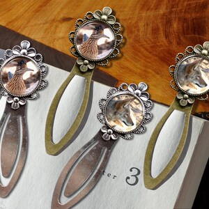 May include: Four metal bookmarks with nature-themed designs. Each bookmark features a circular image of a roadrunner or a wolf, set within a decorative floral frame. The bookmarks are on a book with the number 3 visible.
