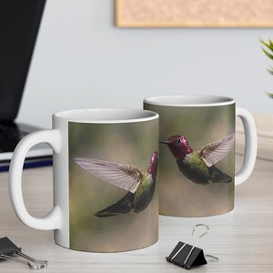 May include: White ceramic coffee mugs featuring a vibrant image of two hummingbirds in flight. The birds have iridescent green bodies, red heads, and outstretched wings, set against a soft, blurred background. The mugs are on a light-colored surface.