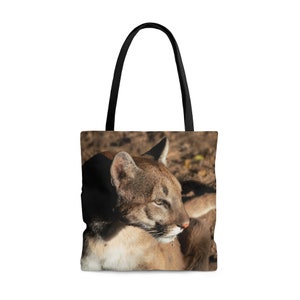May include: A brown and tan cougar with black spots lies on the ground with its head turned to the side. The cougar is looking off to the side with its mouth slightly open. The cougar is lying on a brown and tan background. The image is printed on a black tote bag with black handles.