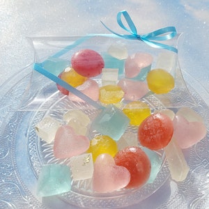 May include: A clear plastic box tied with a blue ribbon filled with colorful heart-shaped and round candies. The candies are on a clear glass plate with a decorative pattern.