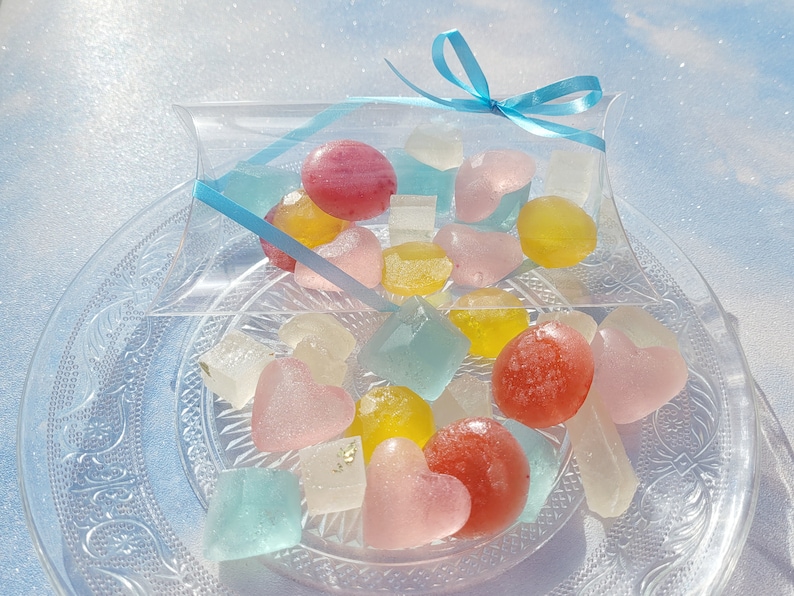May include: A clear plastic box tied with a blue ribbon filled with colorful heart-shaped and round candies. The candies are on a clear glass plate with a decorative pattern.