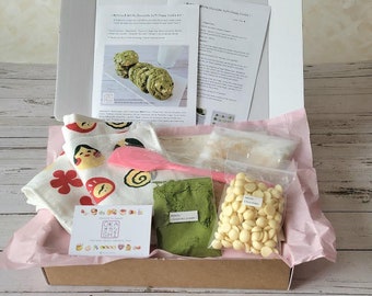 May include: A baking kit for making matcha and white chocolate soft chewy cookies. The kit includes a pink spatula, a bag of white chocolate chips, a bag of matcha powder, two bags of dry ingredients, and three white tea towels with a Japanese-inspired design. The kit also includes instructions for making the cookies.