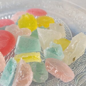 May include: A close-up of a plate of colorful, translucent candy. The candy is in various shapes and sizes, including squares, rectangles, and irregular shapes. The candy is a mix of pink, yellow, green, and clear.