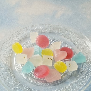 May include: A clear glass plate with a decorative pattern holds a variety of colorful candies. The candies are in various shapes, including hearts, squares, and circles. Some of the candies have a light dusting of gold flakes.