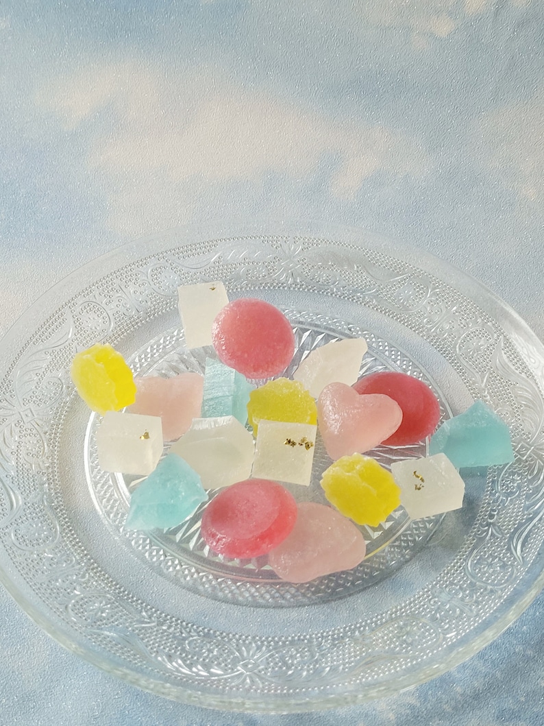 May include: A clear glass plate with a decorative pattern holds a variety of colorful candies. The candies are in various shapes, including hearts, squares, and circles. Some of the candies have a light dusting of gold flakes.