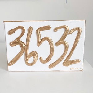 May include: A rectangular white canvas with the numbers "36532" painted in gold. The numbers are in a bold, textured font. The canvas has a gold border and a signature in the bottom right corner.
