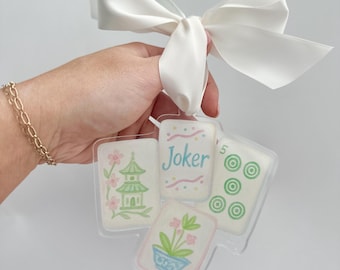 May include: A set of four clear acrylic Mahjong tiles tied with a white satin ribbon. The tiles feature pastel illustrations of a pagoda, flowers, and the word "Joker". The tiles are held together with a white satin ribbon.