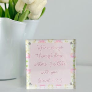May include: A clear acrylic block with the text "When you go through deep waters, I will be with you" and "Isaiah 43:2" in a script font. The block has a pink and green watercolor design. White tulips are in a white vase in the background.
