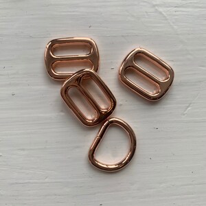 May include: Four rose gold metal hardware pieces for crafting. Three are rectangular with two holes, and one is a D-ring.