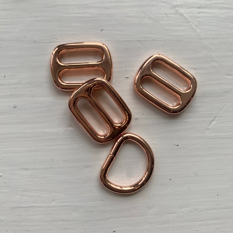May include: Four rose gold metal hardware pieces for crafting. Three are rectangular with two holes, and one is a D-ring.