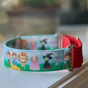 May include: A blue and green dog collar with a red accent. The collar features a printed design of characters from The Wizard of Oz, including Dorothy, the Scarecrow, the Tin Man, the Cowardly Lion, and the Wicked Witch of the West.