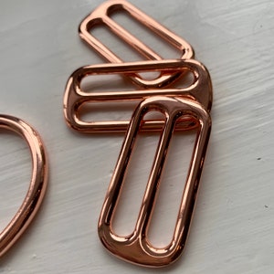 May include: Three rose gold metal slider buckles. The buckles are oval shaped with a smooth, shiny finish.