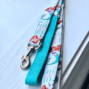 May include: A turquoise blue glitter dog leash with a white leash with a repeating pattern of Ariel from The Little Mermaid and the text "Free as the Sea".