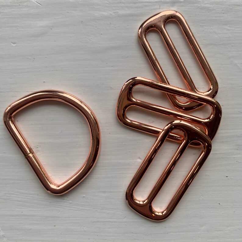 May include: Three rose gold metal slider buckles and one rose gold metal D-ring. The slider buckles are rectangular with two parallel slots. The D-ring is a closed loop.