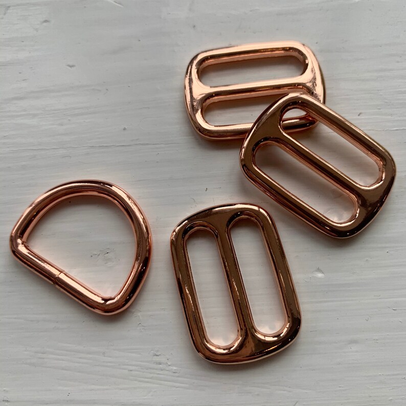 May include: Four rose gold metal slider buckles. Three are rectangular with two parallel slots. One is a D-ring.