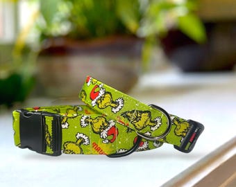 May include: Two green dog collars with a Grinch pattern. The collars feature a black plastic buckle and a metal D-ring. The Grinch is wearing a Santa hat. The background is blurred, with green plants visible.