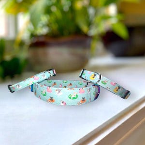 Pode incluir: Um colar e um conjunto de trela para cães azul claro. O colar e a trela apresentam um padrão repetido de cães em boias. O colar tem uma fivela de metal e um anel em D. O fundo está desfocado.