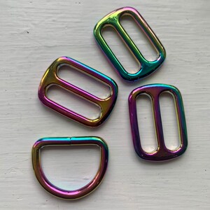 May include: Four iridescent metal slider buckles in various shapes. Two are rectangular with a double-loop design, one is a D-ring, and one is a rectangular buckle with a single loop.