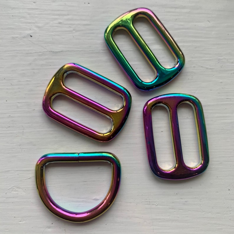 May include: Four iridescent metal slider buckles in various shapes. Two are rectangular with a double-loop design, one is a D-ring, and one is a rectangular buckle with a single loop.