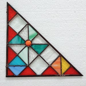 Puede incluir: Un panel triangular de vidriera con un diseño geométrico. El panel presenta piezas de vidrio rojas, azules, verdes, naranjas y amarillas, con un círculo naranja central. El vidrio está enmarcado por metal marrón oscuro.
