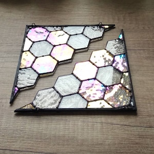 Op de afbeelding: Twee glas-in-loodpanelen met een honingraatontwerp. Het glas heeft iriserende en getextureerde secties in wit, grijs en roze tinten, omlijst door zwarte randen. Elk paneel heeft een kleine lus om op te hangen.