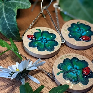 Op de afbeelding: Een paar oorbellen met een ontwerp van een klavertjevier met een lieveheersbeestje op elk klavertje. De klavertjes zijn groen geverfd met blauwe accenten en de lieveheersbeestjes zijn rood met zwarte stippen. De oorbellen zijn van hout en hebben zilveren kettingen.