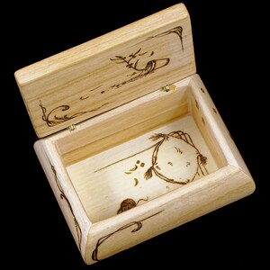 以下が含まれることがあります： 蓋付きの木製の箱で、蓋にはファンシーな生き物と花のモチーフが焼き付けられています。箱は開いており、中は空です。