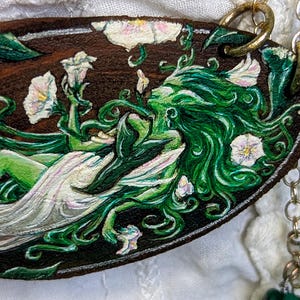 May include: Oval pendant featuring a painted illustration of a woman with green hair and vines, adorned with white flowers. The artwork is set against a dark brown background with gold-colored chain attachments.