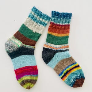 Peut inclure: Une paire de chaussettes tricotées à la main avec un motif rayé de différentes couleurs. Les chaussettes présentent des bandes de bleu, de vert, de marron et de rouge, avec une base bleu clair et un bout multicolore. Les poignets sont bleu foncé.