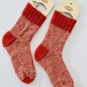 Puede incluir: Un par de calcetines de punto con un patrón de rayas horizontales finas en color naranja quemado y blanco. Los calcetines tienen el puño acanalado, el talón y la puntera en naranja quemado sólido. Cada calcetín está sujeto a una etiqueta de madera clara con la marca OPRY.
