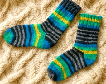 May include: Two colorful knitted socks with horizontal stripes in shades of blue, green, yellow, and gray. The socks are displayed on a cream-colored, textured surface, showcasing a cozy, handmade aesthetic. The socks are designed for warmth and comfort.