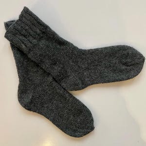 Peut inclure: Une paire de chaussettes tricotées gris foncé. Les chaussettes sont faites d'un fil épais et texturé et ont des poignets côtelés. Les chaussettes sont pliées l'une sur l'autre sur une surface blanche. Les chaussettes semblent chaudes et confortables.