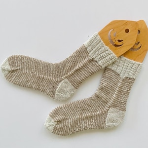May include: A pair of knitted socks featuring a heathered brown and cream color palette. The socks are displayed on light-colored wooden forms that have a cutout design resembling a jack-o'-lantern face at the top of each form.