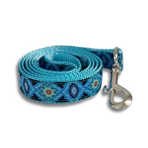 Puede incluir: Una correa para perro turquesa enrollada con un patrón floral decorativo en azul, negro y rojo. La correa tiene un cierre de metal plateado para sujetar al collar.