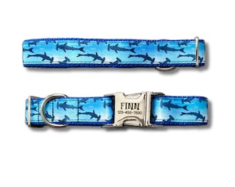 May include: A blue pet collar featuring a repeating pattern of dark blue hammerhead sharks. The collar includes a silver-toned metal side-release buckle engraved with the name FINN and the phone number 123-456-7890, along with a metal D-ring.