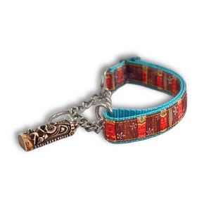 May include: A pet collar featuring a vibrant pattern of red, brown, and orange tiki-style masks on a turquoise blue background. The collar includes a silver-toned metal chain section and a dangling tiki mask charm attached to a metal ring.