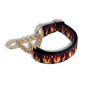 May include: A black fabric dog collar featuring a repeating orange and yellow flame pattern. The collar includes a gold-toned metal chain section with a circular ring attachment and a matching gold-toned buckle mechanism for secure fastening.