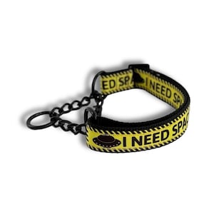 May include: Black dog collar with a yellow band featuring the text "I NEED SPACE" and a UFO graphic. The collar has a black chain and a D-ring for leash attachment. The band has a black and yellow hazard stripe design.