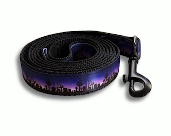 May include: A coiled dog leash featuring a printed pattern of a graveyard silhouette against a purple and blue gradient sky. The leash is made of black webbing and includes a black metal swivel snap hook attached to one end of the strap.