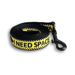 May include: Black dog leash with yellow text that reads "I NEED SPACE". The leash is coiled and has a black metal clip. The yellow text is bordered by a black and white pattern.