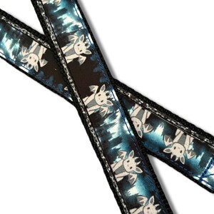 May include: A black fabric strap featuring a repeating pattern of light gray winged creatures with horns. The design includes a blue and black gradient background with vertical streaks. The strap has black stitched edges and a smooth texture.