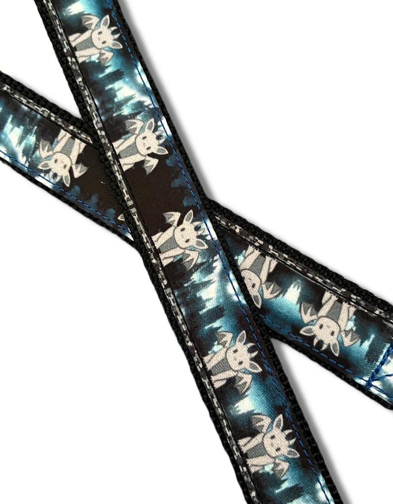 May include: A black fabric strap featuring a repeating pattern of light gray winged creatures with horns. The design includes a blue and black gradient background with vertical streaks. The strap has black stitched edges and a smooth texture.