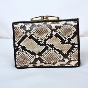 Puede incluir: Bolso de mano cuadrado con un estampado de piel de serpiente en tonos marrón, beige y negro. El bolso tiene un borde negro y un cierre dorado. Es probable que el bolso esté hecho de cuero o un material similar al cuero.