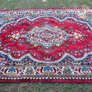 May include: A red rug with a floral pattern and a central medallion. The rug has a dark blue border with a white and gold trim.