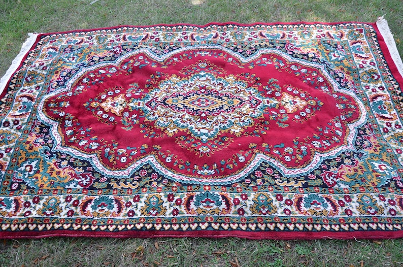 May include: A red rug with a floral pattern and a central medallion. The rug has a dark blue border with a white and gold trim.