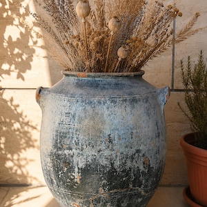 Könnte beinhalten: Eine große, verwitterte blau-graue Keramikvase mit zwei Henkeln, gefüllt mit getrockneten Gräsern und Samenkapseln. Die Vase hat eine abgenutzte Oberfläche, die ihren rustikalen Charme unterstreicht. Ein Terrakottatopf mit einer kleinen Pflanze ist im Hintergrund zu sehen.