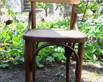 May include: A wooden chair with a curved back and legs. The chair is made of dark brown wood and has a natural finish. The chair is sitting on a dirt ground with green plants in the background.